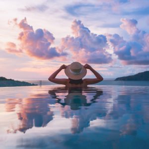 A person in a sunhat relaxes in an infinity pool, facing a scenic view of colorful clouds at sunset over distant hills, with the sky reflected in the water.